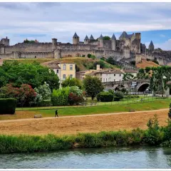 Majestatyczna twierdza Carcassonne góruje nad miastem. W dole zielony park, rzeka i samotny spacerowicz.