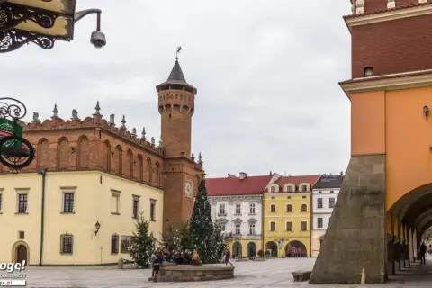 Rynek w Tarnowie zimową porą, z choinką i świątecznymi iluminacjami. W tle charakterystyczna wieża ratuszowa i kolorowe kamieniczki.