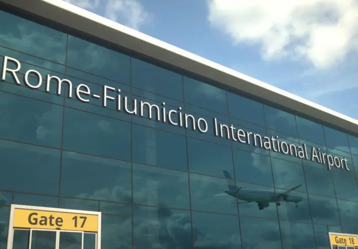 Szklana fasada lotniska w Rzymie, z napisem "Rome-Fiumicino International Airport". W odbiciu widać samolot i chmury.