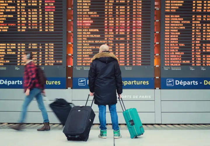 Tablica odlotów na lotnisku z podróżnymi ciągnącymi walizki.