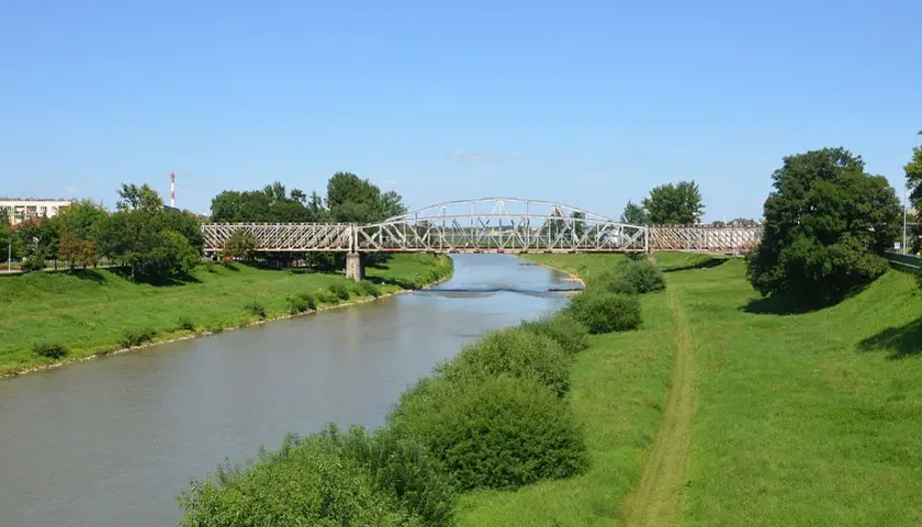 Metalowy most kratownicowy nad rzeką, otoczony zielonymi brzegami i drzewami.
