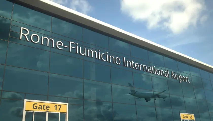 Szklana fasada lotniska w Rzymie, z napisem "Rome-Fiumicino International Airport". W odbiciu widać samolot i chmury.