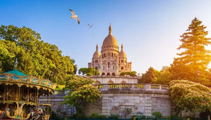 Bajkowy karuzela na tle bazyliki Sacré-Cœur na Montmartre. Mewy szybują na błękitnym niebie.