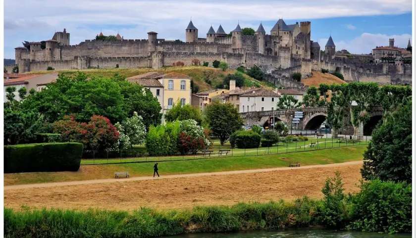 Majestatyczna twierdza Carcassonne góruje nad miastem. W dole zielony park, rzeka i samotny spacerowicz.