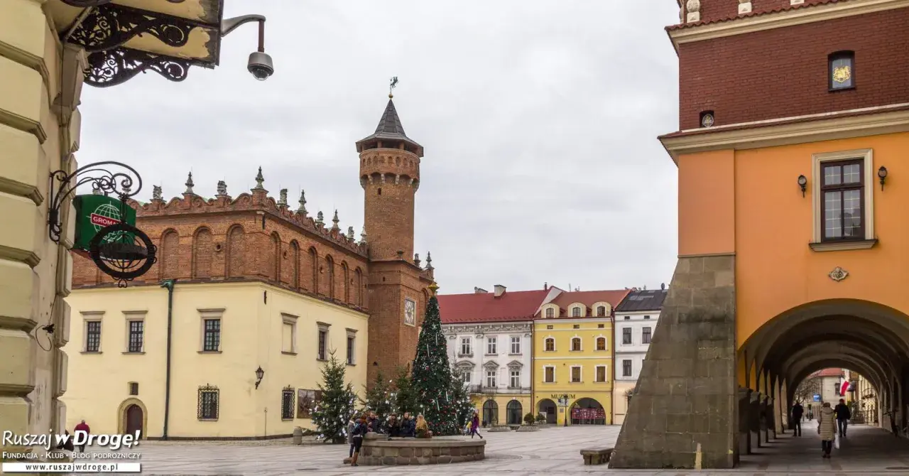 Rynek w Tarnowie zimową porą, z choinką i świątecznymi iluminacjami. W tle charakterystyczna wieża ratuszowa i kolorowe kamieniczki.