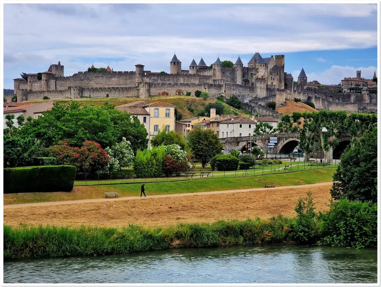 Majestatyczna twierdza Carcassonne góruje nad miastem. W dole zielony park, rzeka i samotny spacerowicz.